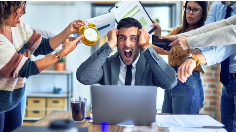 A man at a desk holding his head while colleagues crowd in with a clock, a phone and paperwork