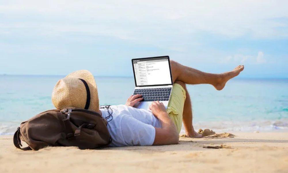 A man lying on a beach working on a laptop