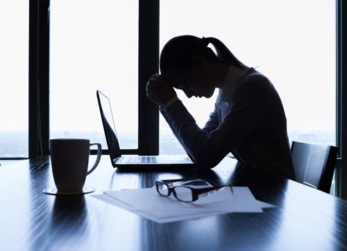 A person sitting at a desk with their head in their hands, silhouetted against an office window